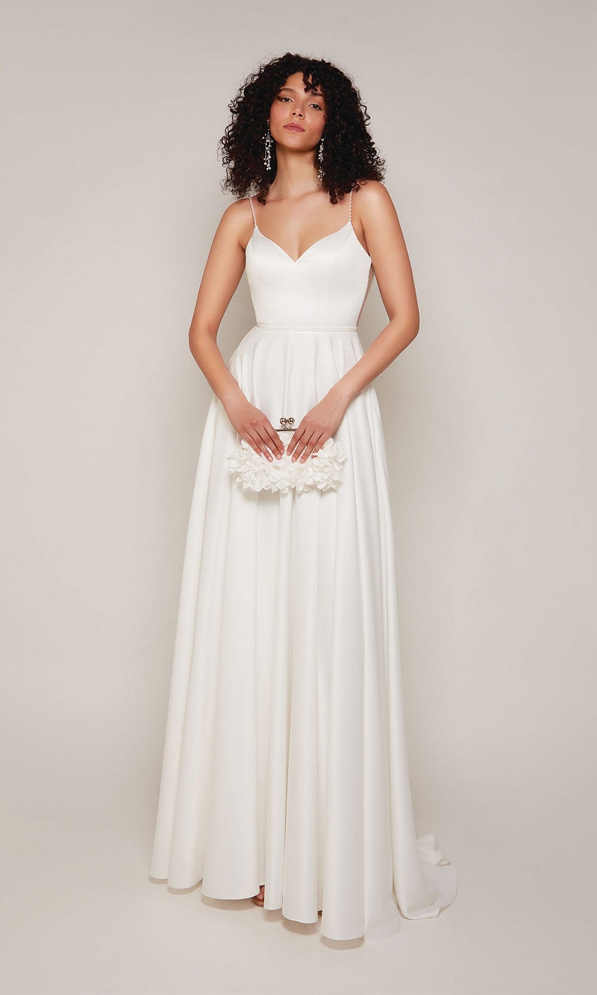 A woman in the Alyce 7099 long white dress with beaded spaghetti straps holds a small white clutch, standing against a plain light background.
